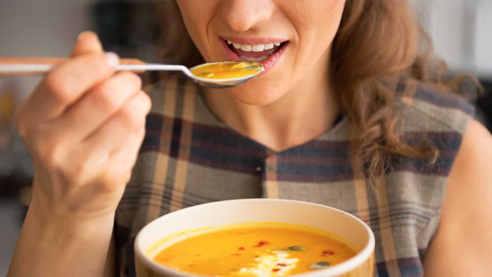 A person is holding a spoonful of orange soup close to their mouth, ready to take a bite. They are also holding a bowl filled with the same soup, which appears to be pumpkin or squash soup, garnished with seeds and a drizzle of cream.