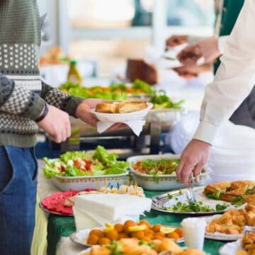 People are serving themselves food from a buffet table. The spread includes salads, bread, and fried appetizers. Some individuals are holding plates, and others are dressed in formal attire, suggesting a catered event.