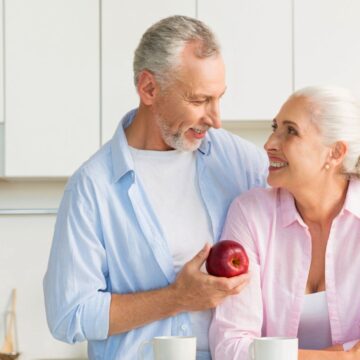 An older couple stands in a kitchen. The man, holding an apple, has his arm around the woman's shoulder. Both are smiling at each other. Behind them are kitchen utensils, a potted plant, and shelves with bowls and fruit.