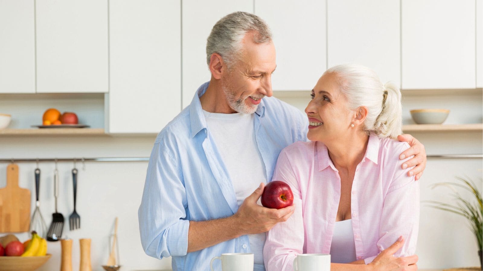 An older couple stands in a kitchen. The man, holding an apple, has his arm around the woman's shoulder. Both are smiling at each other. Behind them are kitchen utensils, a potted plant, and shelves with bowls and fruit.