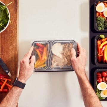 A person arranges meal prep containers on a table. The containers hold sliced chicken, vegetables, and brown rice. Surrounding the containers are a knife, spinach, broccoli, an avocado, and sliced peppers.