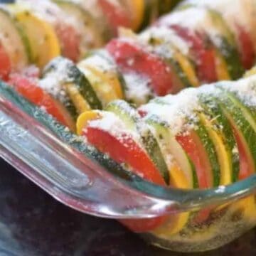 A glass baking dish filled with slices of tomato, zucchini, and yellow squash arranged upright in rows, sprinkled with grated cheese or breadcrumbs on top, ready for baking.