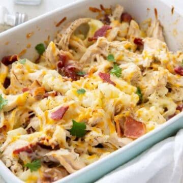 A white baking dish filled with a casserole made of shredded chicken, melted cheese, bacon pieces, and garnished with parsley. A white cloth and a pepper shaker are nearby.