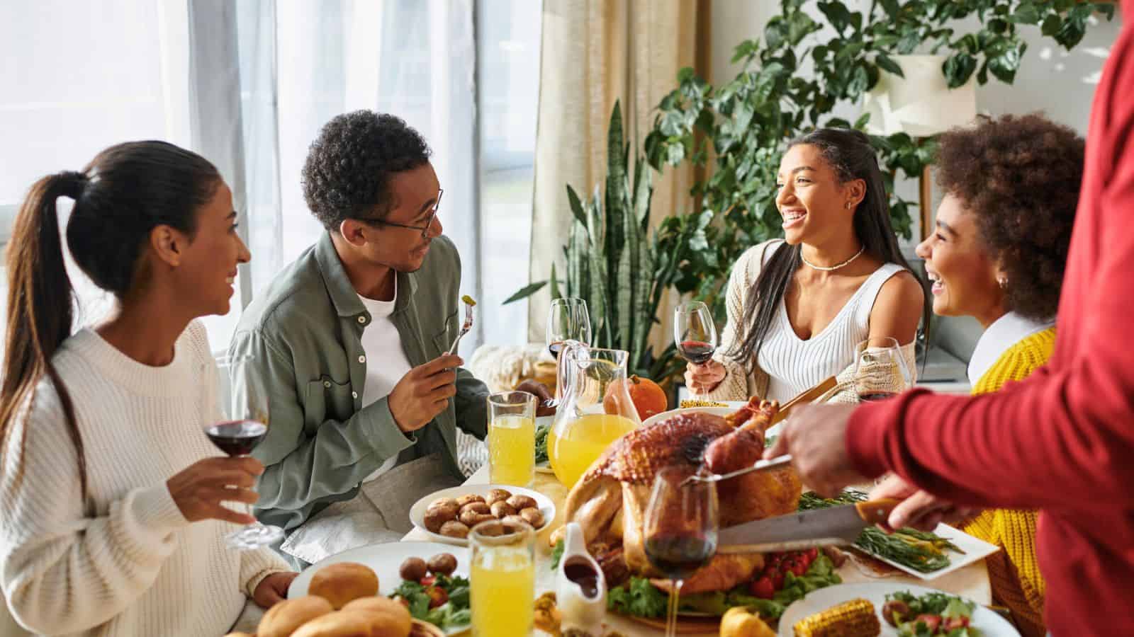 A group of people sitting at a dining table, enjoying a meal together. The table is set with a roast turkey, bread rolls, and various side dishes. They are holding drinks and engaging in conversation, smiling. A leafy plant is in the background.