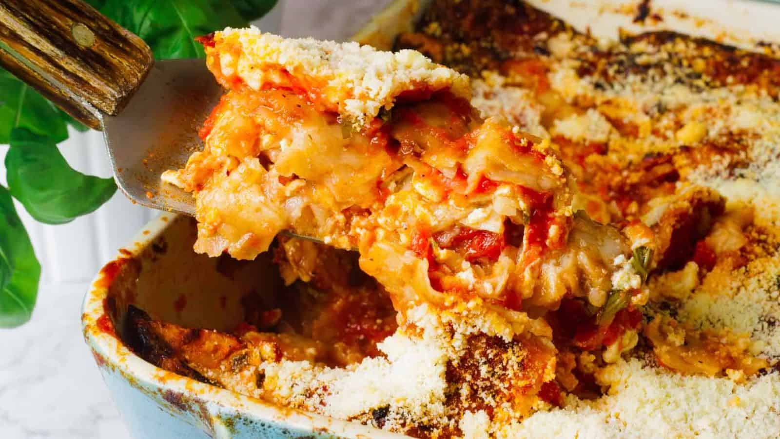 A slice of baked lasagna is being lifted from a casserole dish. The lasagna layers are visible, with melted cheese, tomato sauce, and pasta. The dish is sprinkled with grated cheese. A leafy green plant is partially visible in the background.