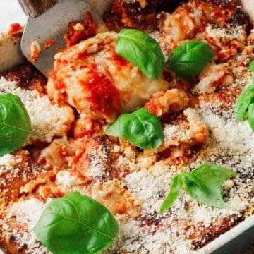 A baked lasagna in a rectangular dish, topped with tomato sauce, basil leaves, and grated cheese. A spatula is lifting a portion from the dish, revealing layers of pasta and filling. The dish is on a light countertop.