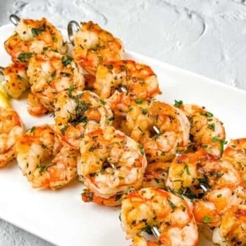 A white rectangular plate with grilled shrimp skewers garnished with herbs, placed next to two lemon wedges on a textured light gray surface.