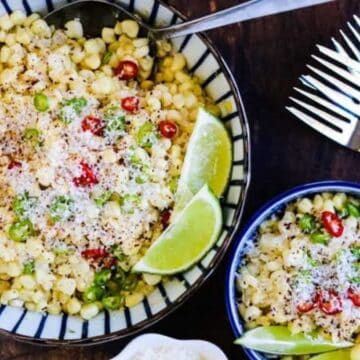 Two bowls of Mexican street corn salad topped with grated cheese, sliced red and green chili peppers, and lime wedges, set on a wooden surface next to a small bowl of cheese and several forks.
