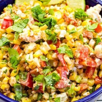 A blue bowl filled with Mexican street corn salad, featuring corn, diced red peppers, cheese, green onions, cilantro, and slices of avocado on top. The bowl is placed next to a floral-patterned cloth.