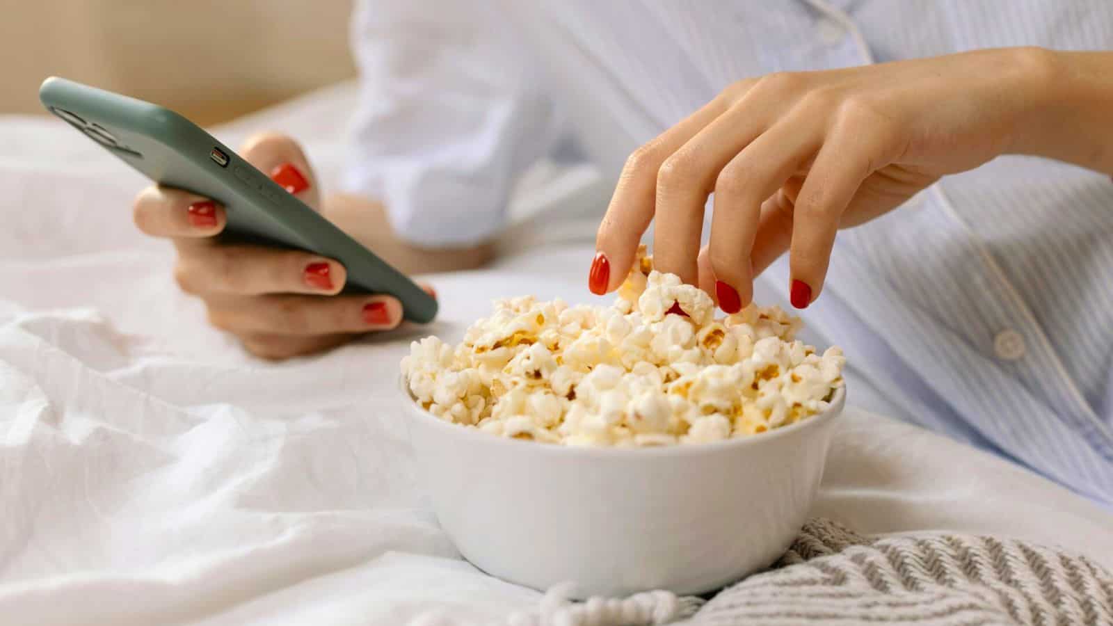A person with red nail polish holds a smartphone in one hand while reaching into a white bowl of popcorn with the other. The scene is set on a comfortable, light-colored bedspread.