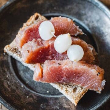 A slice of brown bread topped with three pieces of cured fish and three small white onions on a round, dark plate. The plate is placed on a wooden surface.