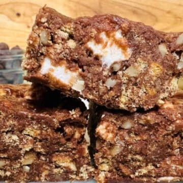 Three stacked chocolate dessert bars with visible nuts, mini marshmallows, and crumbly pieces evoke the charm of classic desserts. A jar of chocolate chips and a jar of marshmallows are blurred in the background.