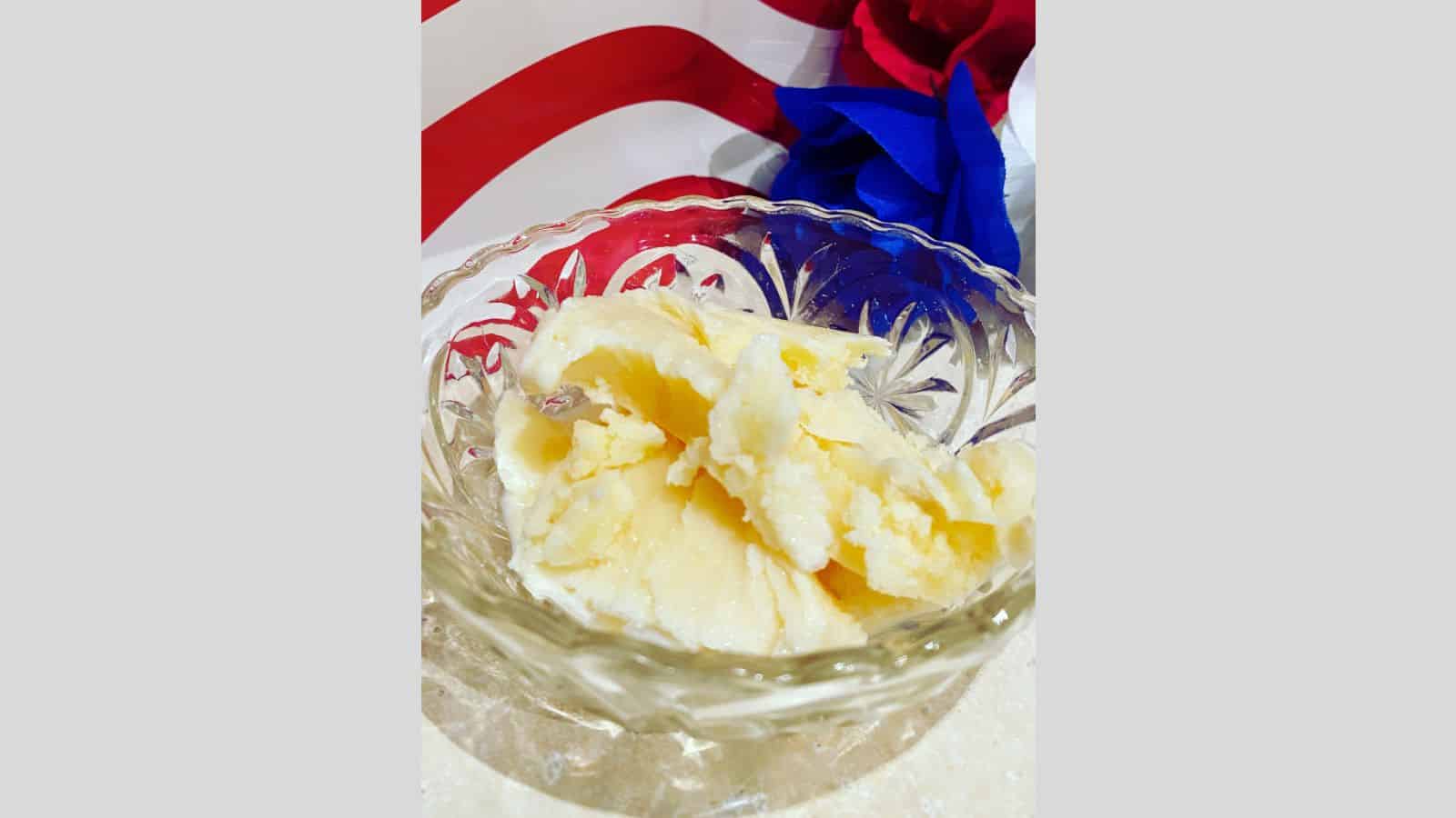 A clear glass bowl with several scoops of vanilla ice cream sits on a light-colored surface. A red, white, and blue background is partially visible behind the bowl, evoking the charm of classic desserts and nostalgic sweets.