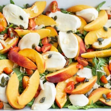A salad in a white dish with sliced peaches, cherry tomatoes, mozzarella cheese, arugula, and pumpkin seeds.