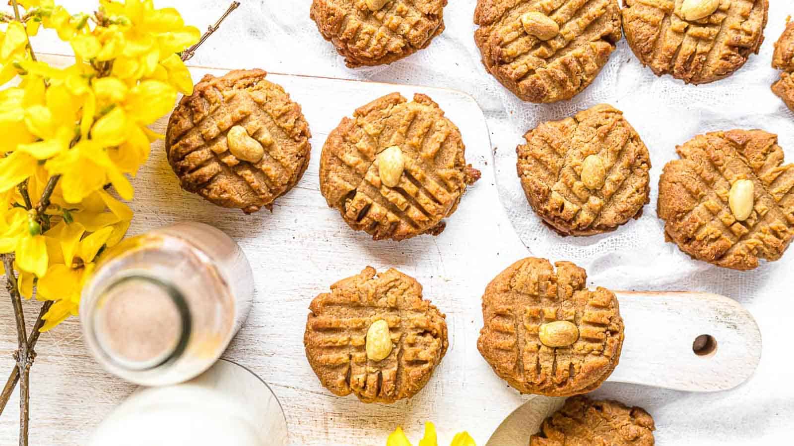 A group of homemade peanut butter cookies with crisscross fork marks and peanuts on top are arranged on a white wooden board, next to a bottle of milk and yellow flowers, on a white surface.