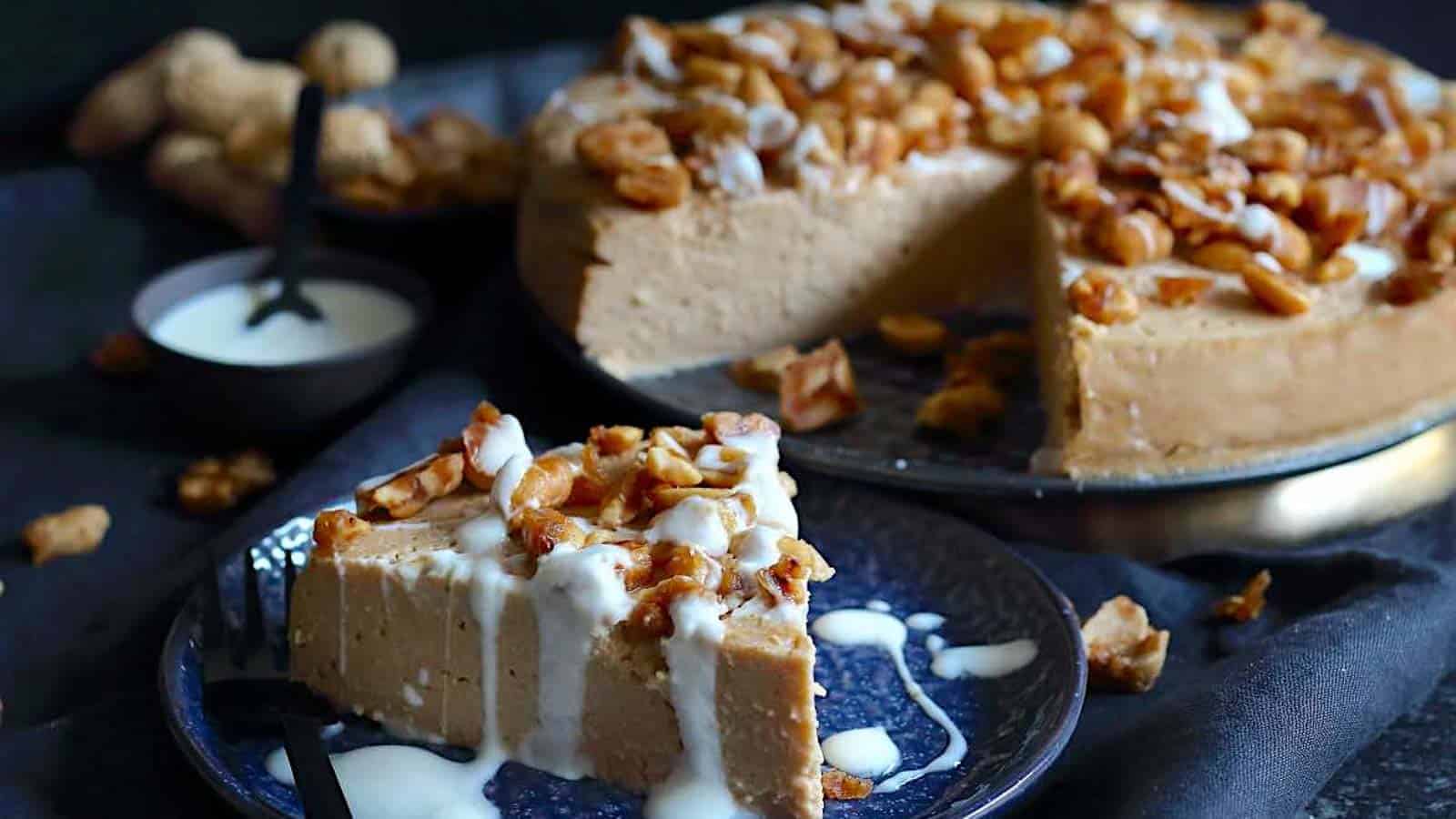 A slice of creamy cheesecake topped with roasted nuts sits on a plate with a drizzle of white sauce, next to the remaining cheesecake topped with the same nuts. A small bowl of sauce and a fork are also visible.