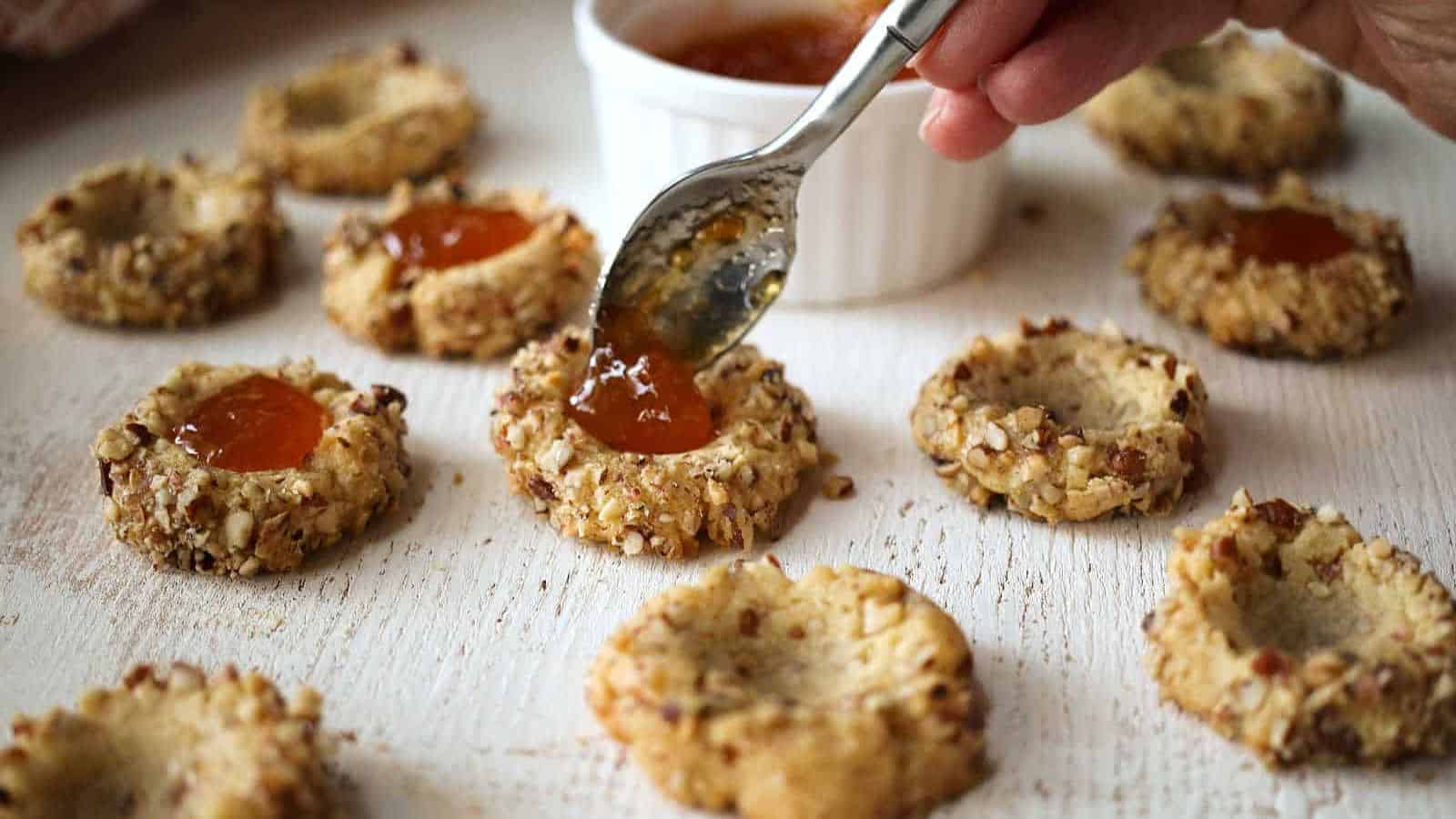 A hand uses a spoon to fill thumbprint cookies with orange jam. These classic desserts are coated with chopped nuts and arranged on a white surface, with a small white bowl of jam in the background.