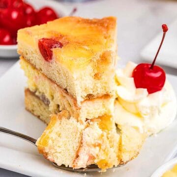 A slice of layered pineapple upside-down cake with whipped cream and a cherry on a white plate, next to a fork. A bowl of cherries and another plate of dessert are in the background.