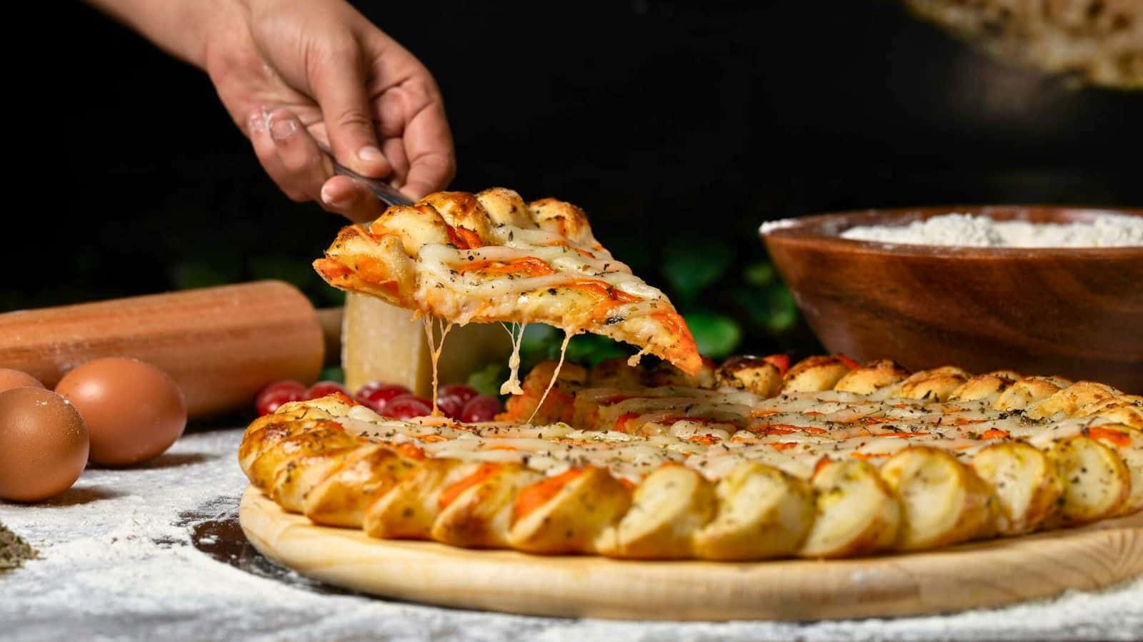 A hand lifts a slice of a cheese pizza with a thick, twisted crust from a wooden board. Ingredients like eggs, flour, tomatoes, and a rolling pin are visible around the pizza. The cheese stretches as the slice is pulled up.