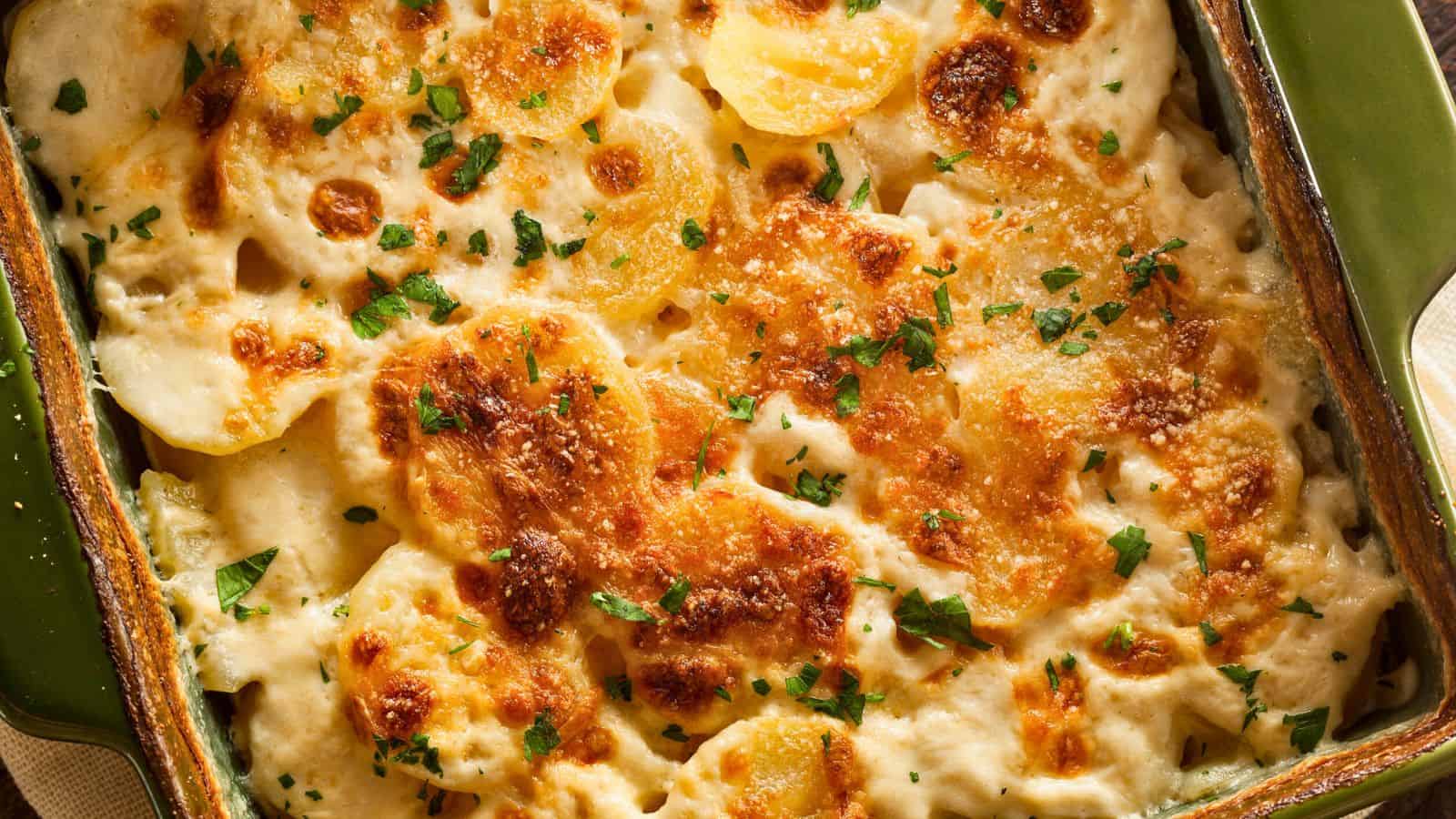 A close-up of a baked potato gratin in a green rectangular dish. The top layer is golden brown with melted cheese and garnished with chopped parsley. The surface shows slices of potatoes in a creamy sauce.