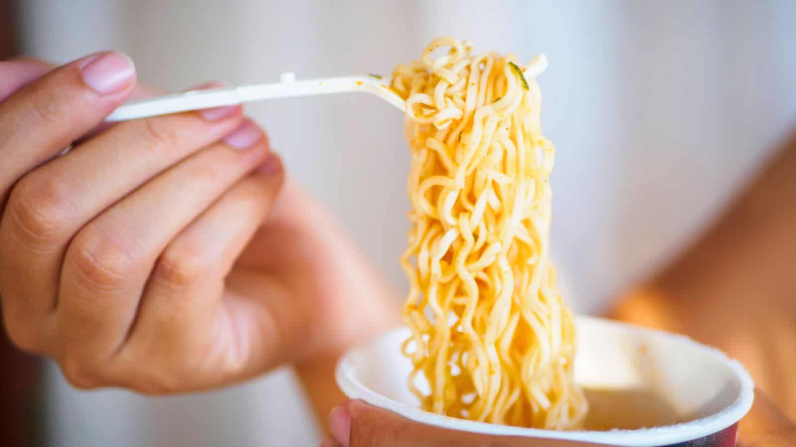 A person holds a cup of instant noodles with one hand while using a fork to lift some noodles out. The focus is on the noodles and the hands, with a blurred background.
