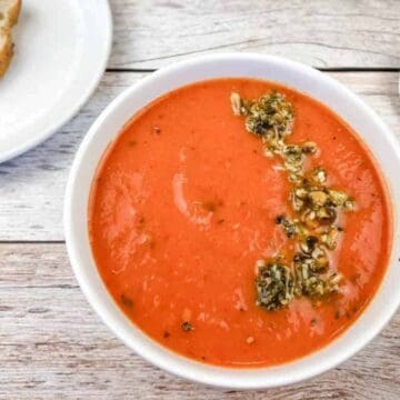 A bowl of creamy tomato soup topped with fresh herbs and seeds sits on a wooden surface, evoking Grandma's kitchen. A stainless steel spoon lies beside it, with a small plate of sliced bread in the background—thanks to timeless recipes that warm the soul.