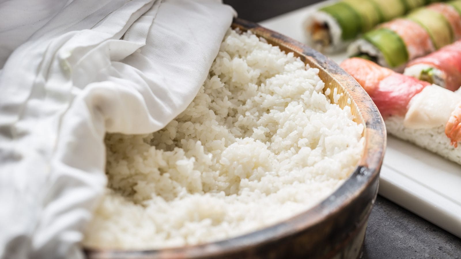 A wooden bowl filled with cooked white rice is partially covered by a white cloth. In the background, assorted sushi rolls are placed on a white tray.