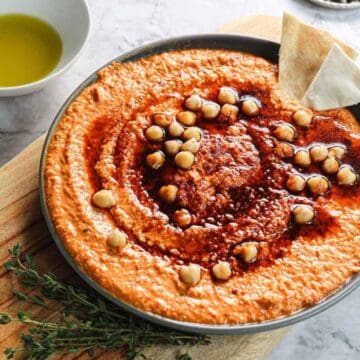 A bowl of creamy hummus topped with chickpeas and a red sauce, placed on a wooden board. Pita bread pieces and a small bowl of olive oil are nearby on a marble surface.