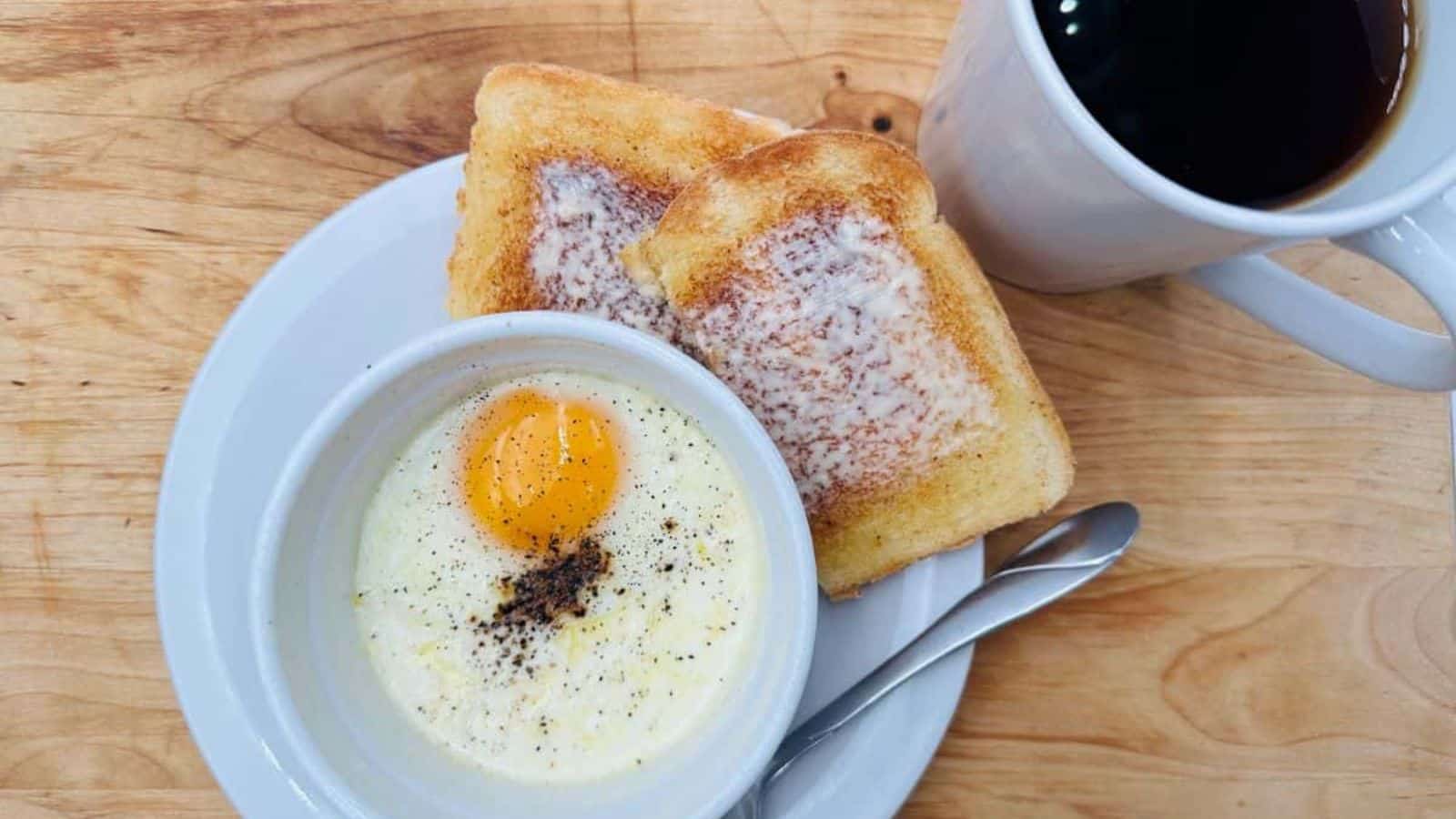 A breakfast setting showcasing classic recipes: a soft-boiled egg topped with pepper in a white bowl, alongside two slices of buttered toast on a white plate. A metal spoon rests by the plate, and a cup of black coffee adorns the wooden table.