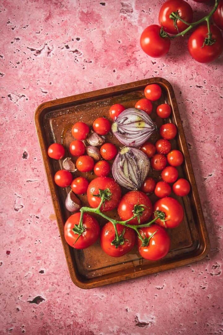 A baking tray with whole tomatoes on the vine, cherry tomatoes, halved red onion, and garlic cloves sits on a pink textured surface—perfect ingredients for salsa or even baked feta in Mexican salsa. More vine tomatoes rest nearby in the corner.