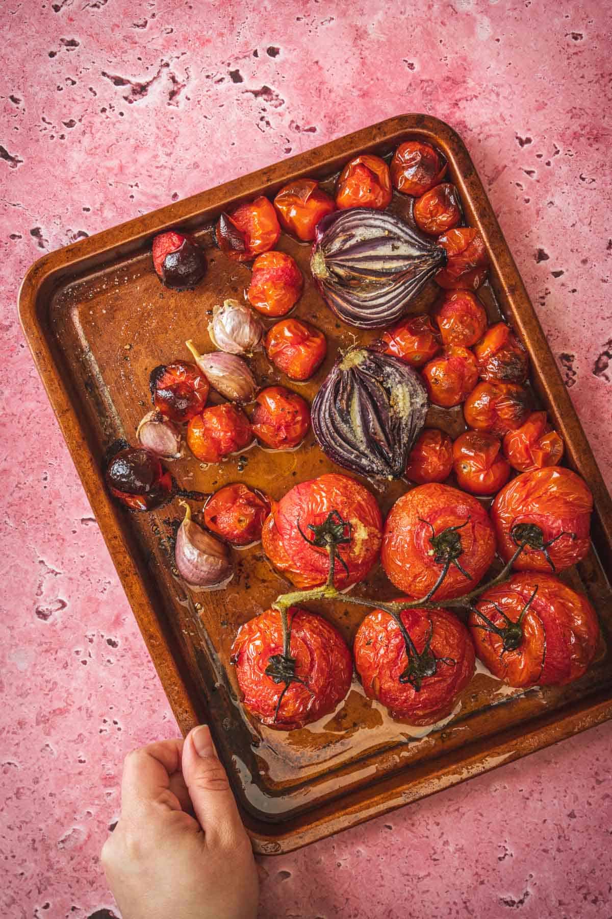 A hand holds a baking tray filled with roasted tomatoes, cherry tomatoes, red onion halves, and garlic cloves—perfect for making a smoky salsa or pairing with baked feta in Mexican salsa. The veggies are slightly charred and glistening on a pink marbled surface.