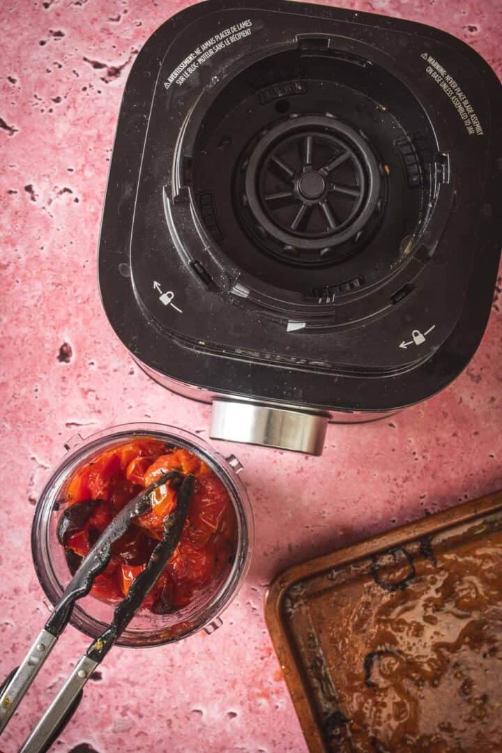 A blender base sits on a pink countertop next to an open blender cup filled with roasted tomatoes for salsa, with metal tongs resting on the cup and a used baking tray nearby—perfect for making baked feta in Mexican salsa.