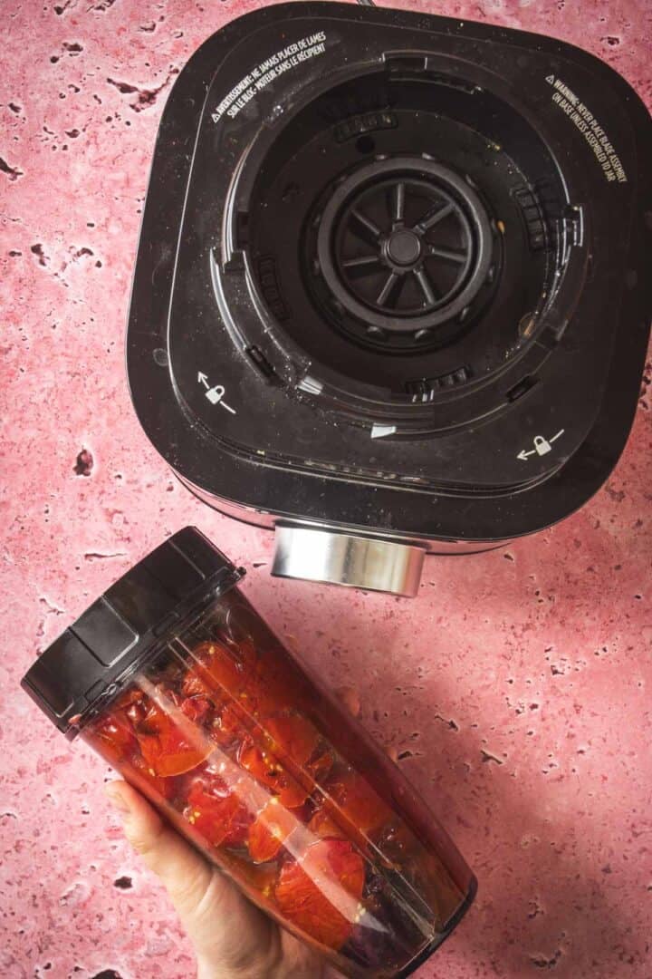 A hand holds a blender cup filled with chopped tomatoes and veggies for salsa, positioned next to the base of a black countertop blender on a pink marbled surface—perfect for whipping up fresh dips or baked feta in Mexican salsa.