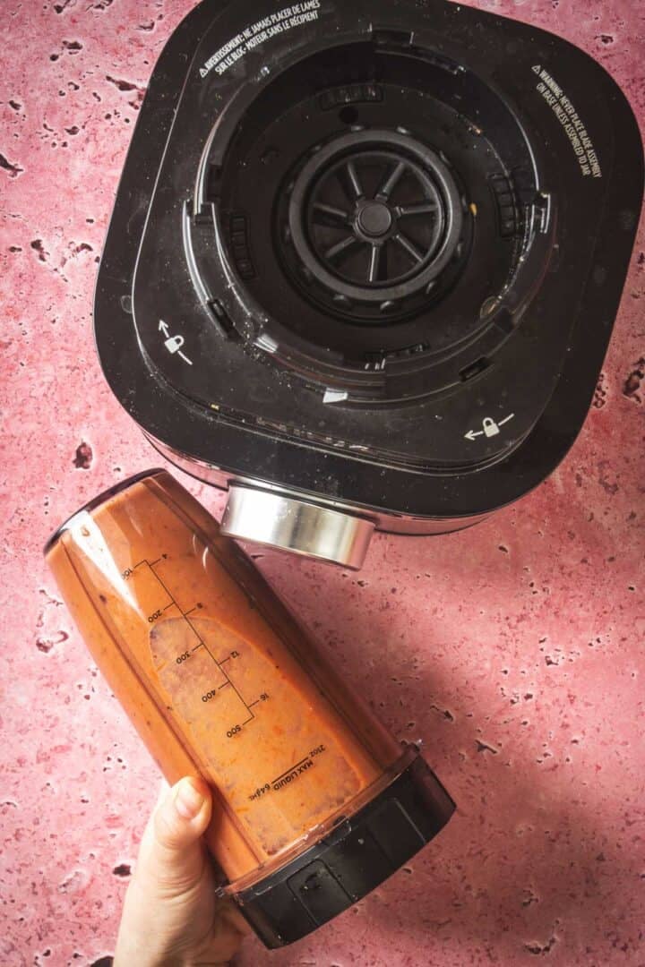 A hand holds a blender cup filled with an orange-brown salsa mixture next to the base of a blender, all placed on a pink countertop. Measurement markings are visible on the cup, perfect for whipping up Baked Feta in Mexican Salsa.