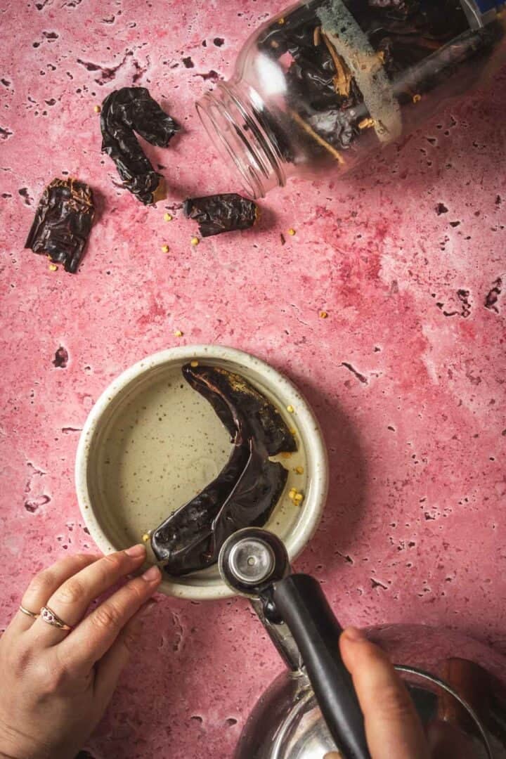 A person holds tongs and places a dried chili pepper into a bowl on a pink textured surface, preparing ingredients for baked feta in Mexican salsa. A jar of chilies is tipped over, with pieces scattered around, ready to spice up the salsa.
