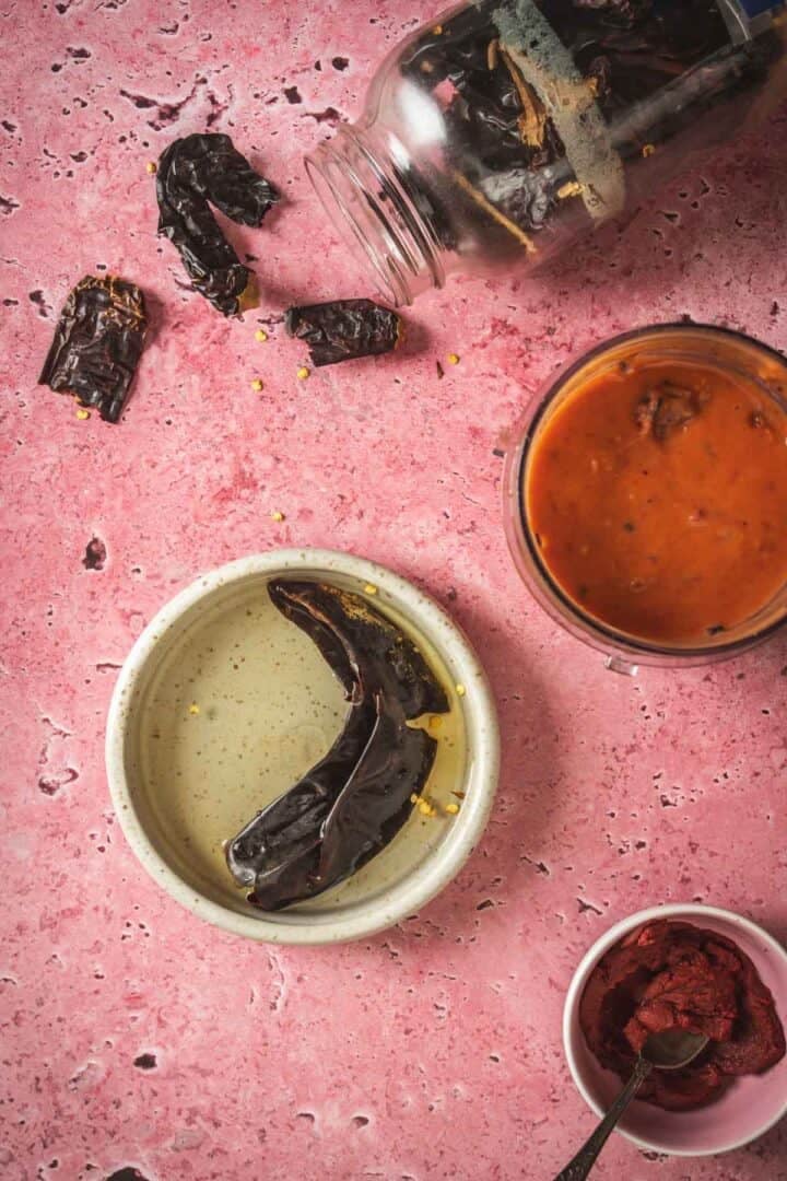A jar of dried chiles is spilled on a pink surface, next to a bowl of chiles soaking in liquid, a glass with red salsa, and a small bowl of tomato paste—perfect ingredients for making baked feta in Mexican salsa.
