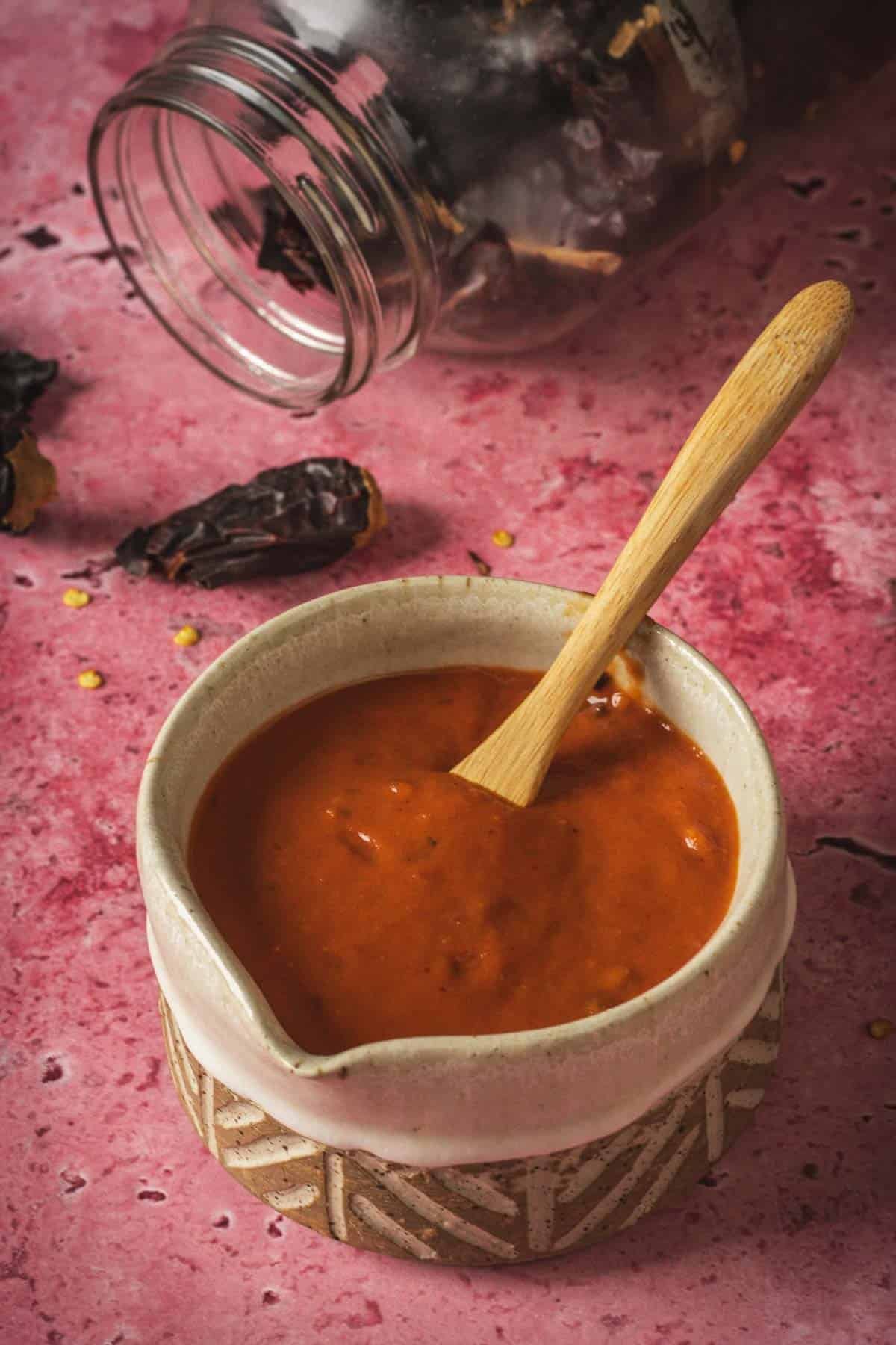 A ceramic bowl filled with vibrant salsa and a wooden spoon sits on a patterned coaster. In the background, a glass jar lies on its side with dried chilies spilling out, evoking flavors of baked feta in Mexican salsa.