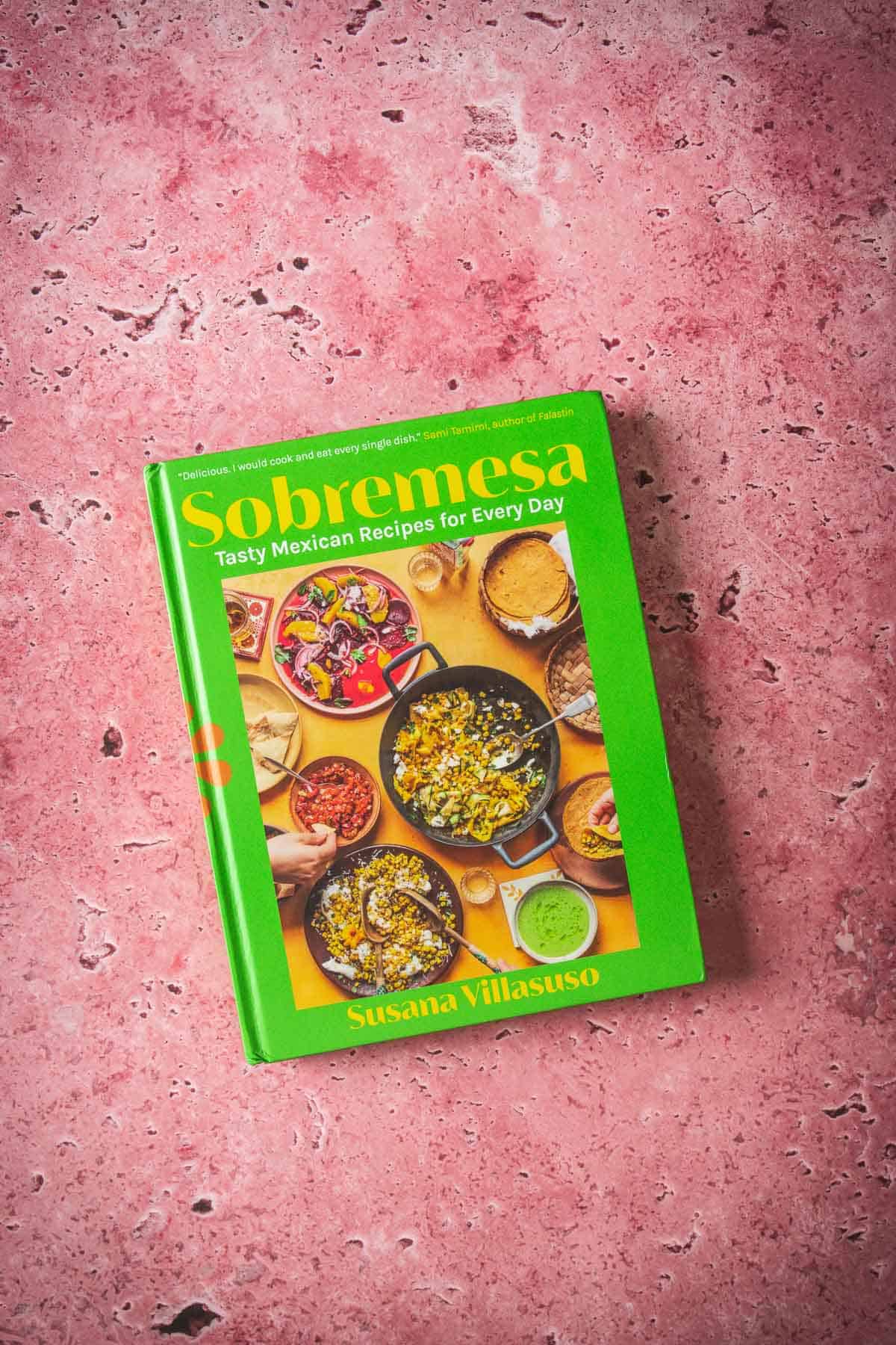 A green hardcover cookbook titled "Sobremesa: Tasty Mexican Recipes for Every Day" by Susana Villasuso rests on a pink, marbled surface. The cover displays overhead photos of Mexican dishes, including Salsa and Baked Feta in Mexican Salsa.
