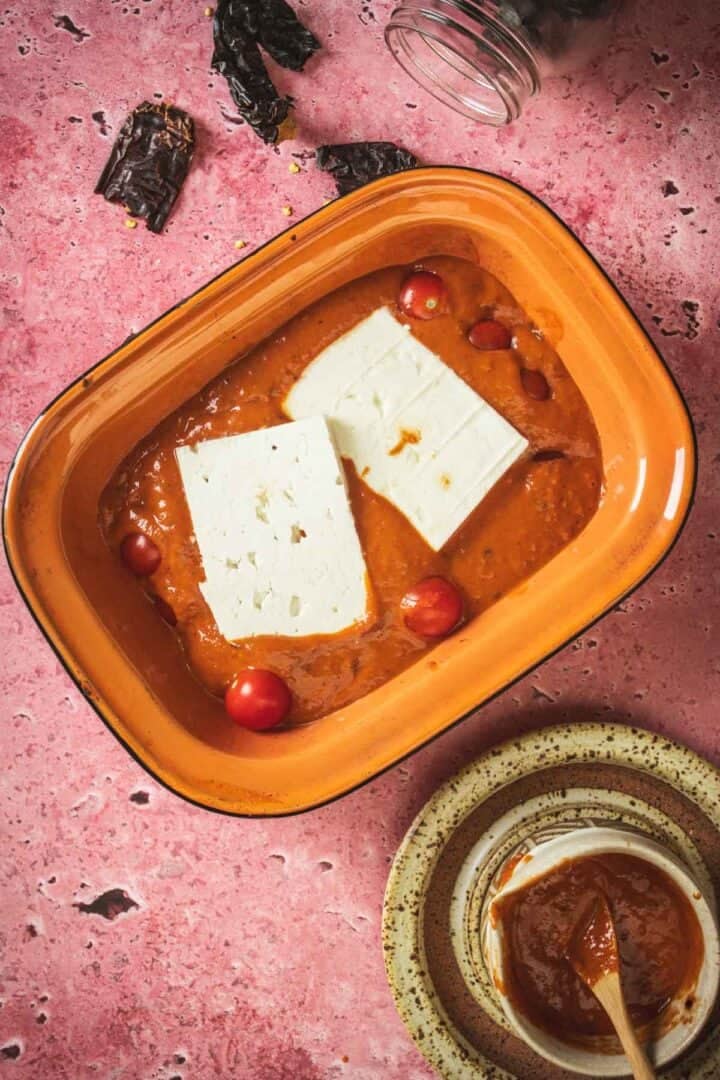 An orange rectangular baking dish holds two blocks of white cheese for a Baked Feta in Mexican Salsa, with cherry tomatoes and red salsa. A bowl of extra salsa, dried peppers, and a glass jar sit nearby on a pink surface.