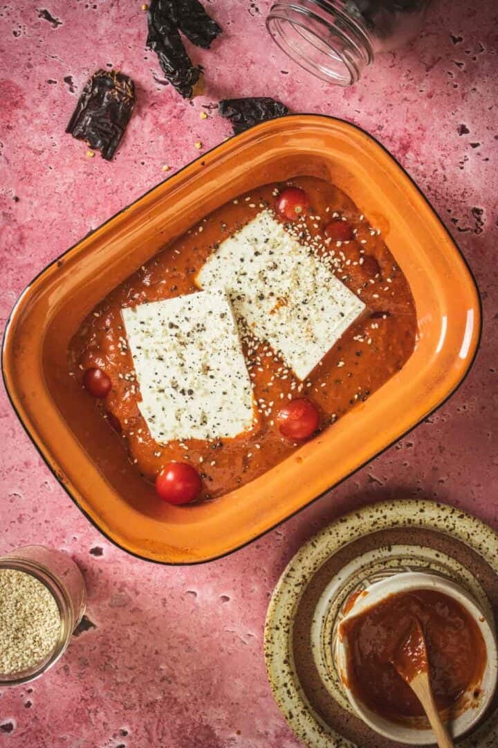 A rectangular orange baking dish with two blocks of baked feta in Mexican salsa, cherry tomatoes, and sprinkled seeds on top, placed on a pink surface with a bowl of salsa and scattered dried chiles nearby.