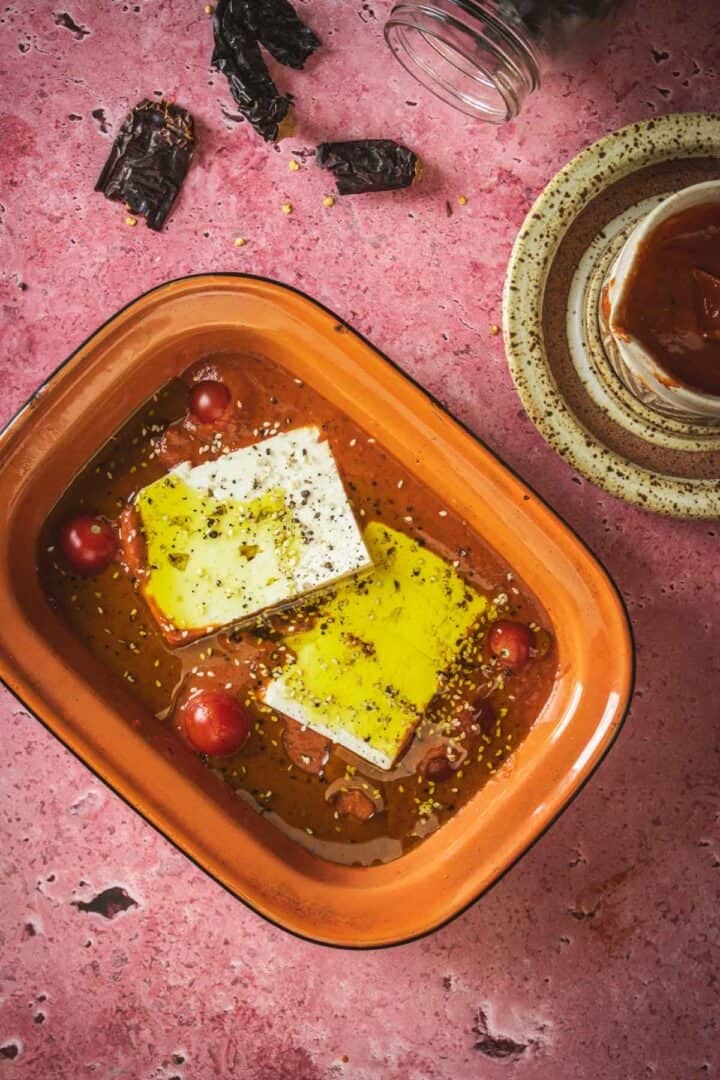 A rectangular orange baking dish holds two blocks of feta cheese, cherry tomatoes, olive oil, and spices in a zesty Mexican salsa, set on a pink textured surface with a jar, a small plate, and dried chilies nearby.