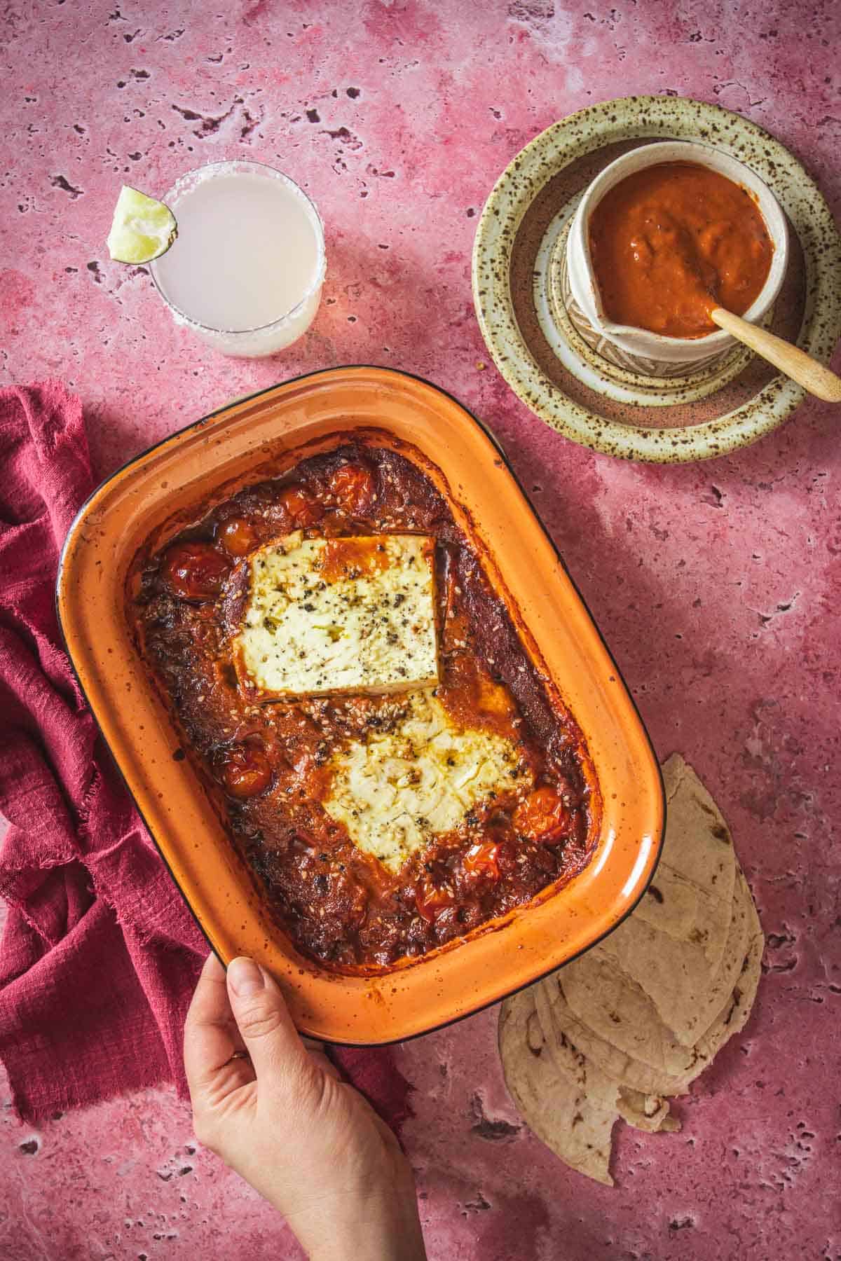 A hand holds a rectangular baking dish with baked feta in Mexican salsa and tomatoes. Nearby are folded tortillas, a bowl of salsa with a spoon, a drink with a lime wedge, and a red cloth on a pink surface.
