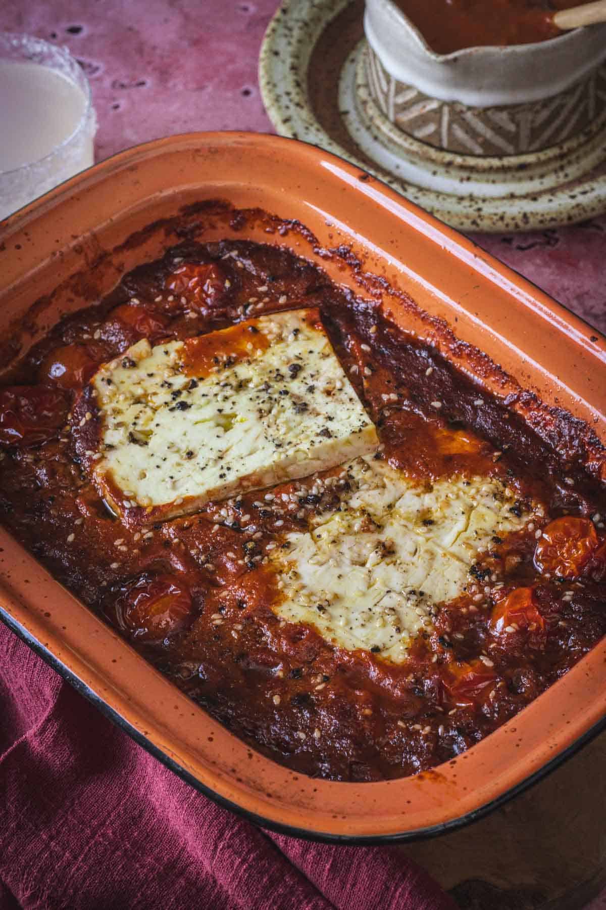 A baked dish with a large block of feta cheese on top of roasted cherry tomatoes and rich Mexican salsa, seasoned with herbs and black pepper, in an orange-brown baking pan on a red tablecloth.