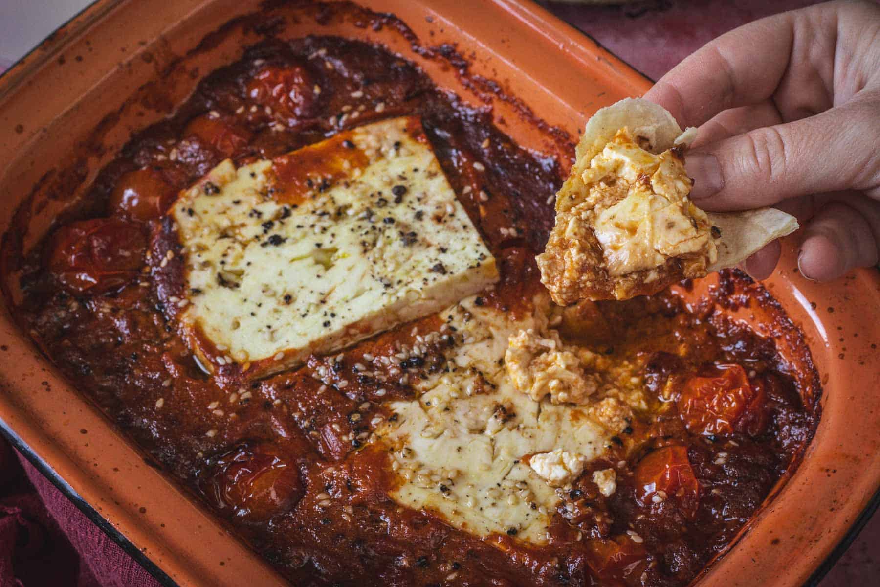 A hand holds a piece of bread scooping Baked Feta in Mexican Salsa from a pan with roasted cherry tomatoes and sauce. The feta is seasoned with herbs, black pepper, and zesty salsa flavors.