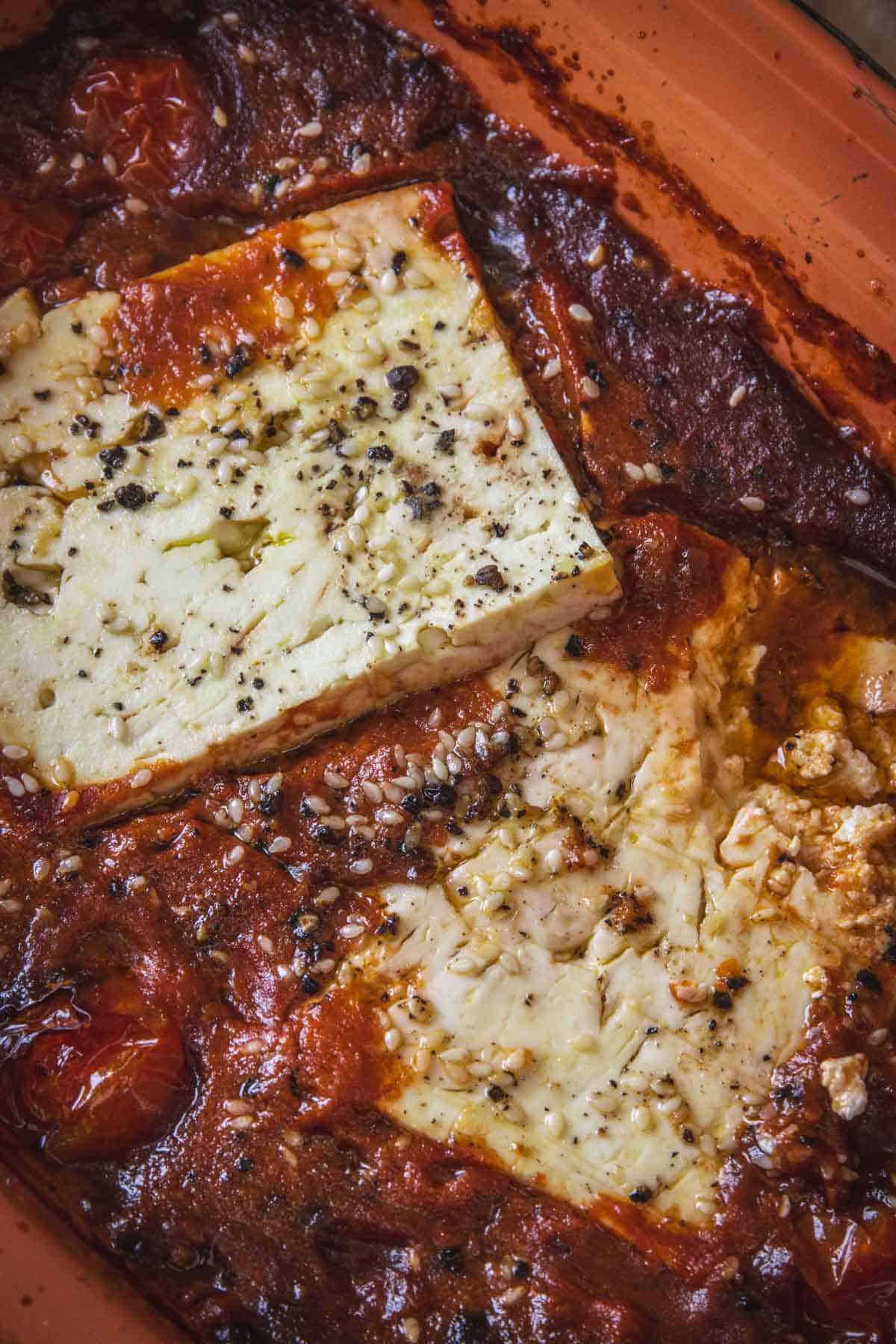 Rectangular blocks of feta cheese seasoned with cracked black pepper sit on top of a baked tomato salsa in a dish. The edges of the Mexican salsa are caramelized, surrounding the creamy, oven-baked feta for a flavorful twist.