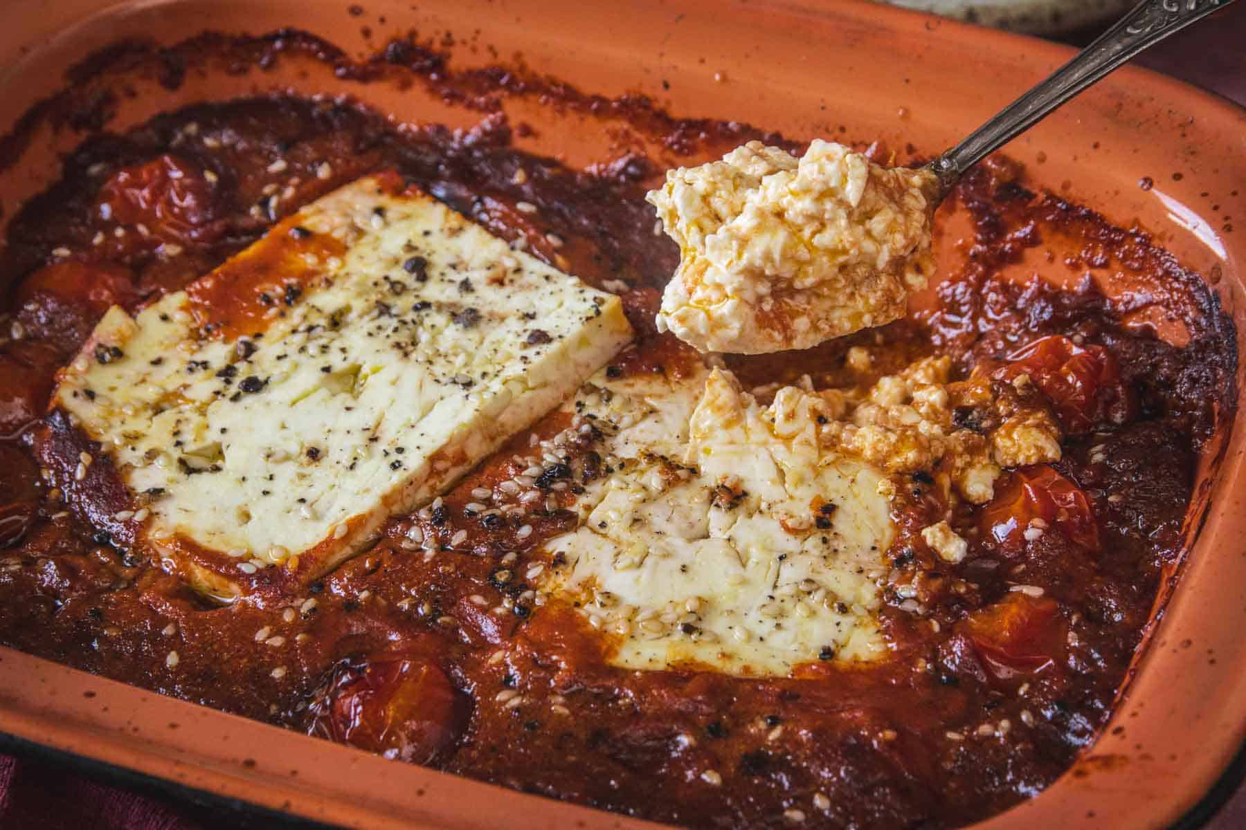 A close-up of Baked Feta in Mexican Salsa, featuring two large feta blocks nestled on roasted cherry tomatoes and rich salsa, seasoned with herbs; a spoon scoops out a flavorful portion of feta and sauce.