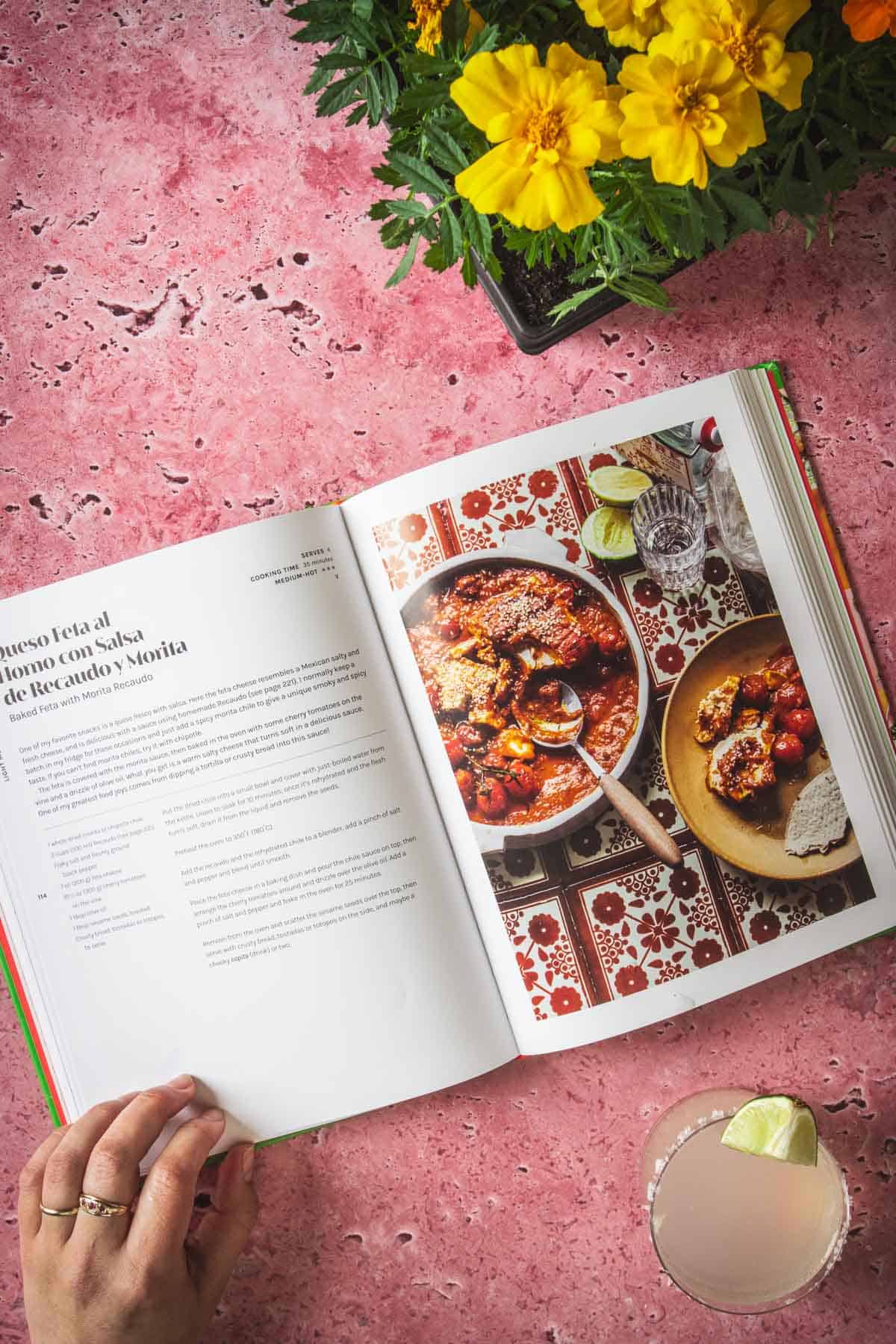 An open cookbook lies on a pink marble surface next to a glass of a light-colored drink with a lime wedge and a yellow flower plant. The recipe features Baked Feta in Mexican Salsa, with an enticing photo of the dish in a pan.