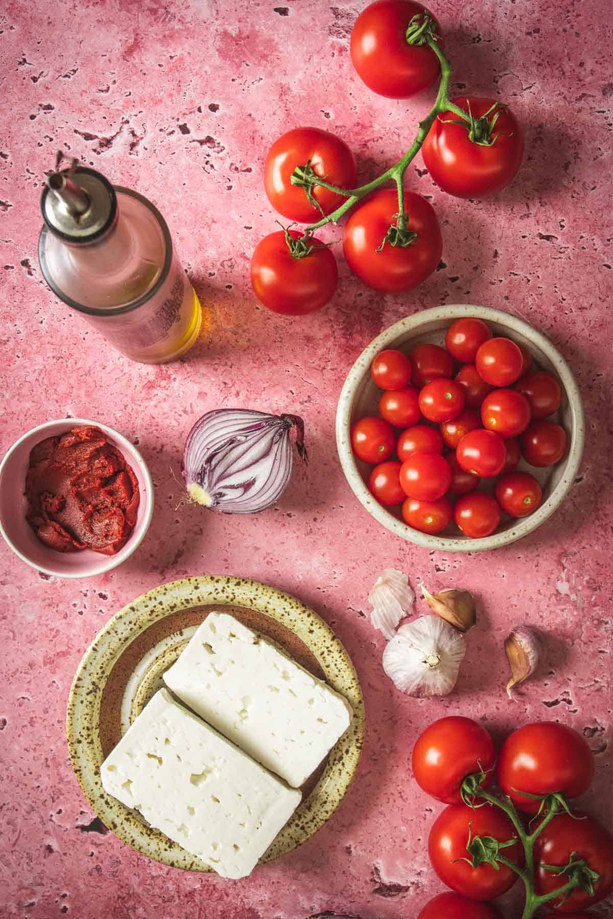 A flat lay of Mediterranean ingredients on a pink surface: olive oil, vine tomatoes, cherry tomatoes, red onion, tomato paste, garlic cloves, and feta cheese—perfect for creating a flavorful Baked Feta in Mexican Salsa twist.