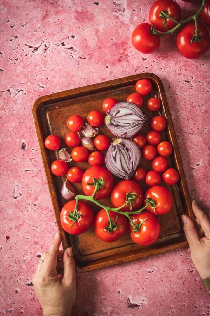 A person holds a wooden tray filled with various red tomatoes, whole garlic bulbs, and halved red onions—perfect ingredients for salsa or baked feta in Mexican salsa—on a pink stone surface, with more vine tomatoes in the top right corner.