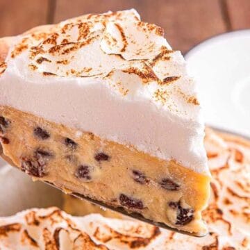 A slice of pie with a golden-brown meringue topping and a filling containing chocolate chips is being lifted from the whole pie, which is on a wooden table next to a white plate and forks.